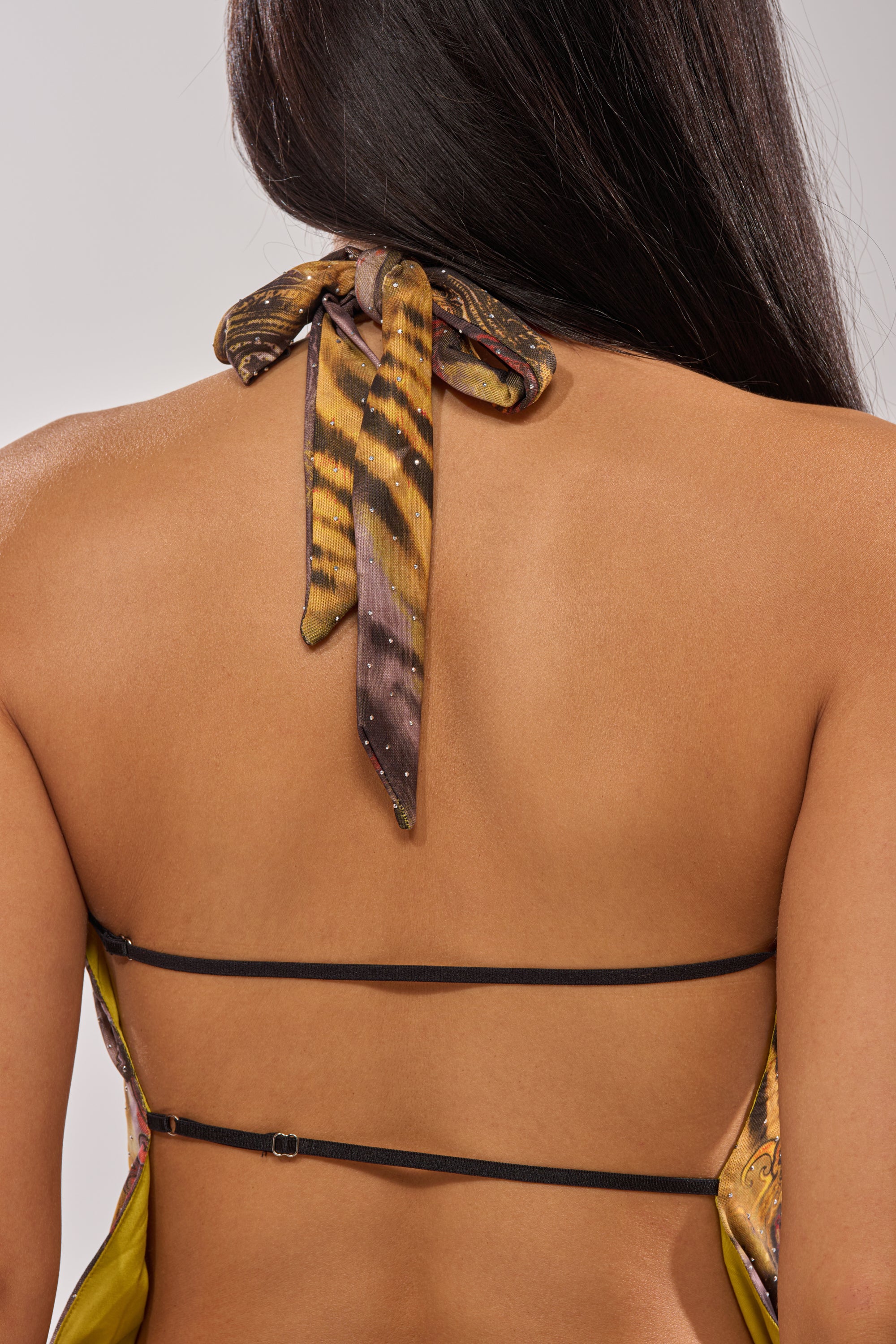 A close-up of a woman's back showcases the JAGUAR GLOW MINI DRESS, with a jungle-inspired print, brown and yellow neck tie, and two thin black straps. Her long dark hair cascades over one shoulder. 8