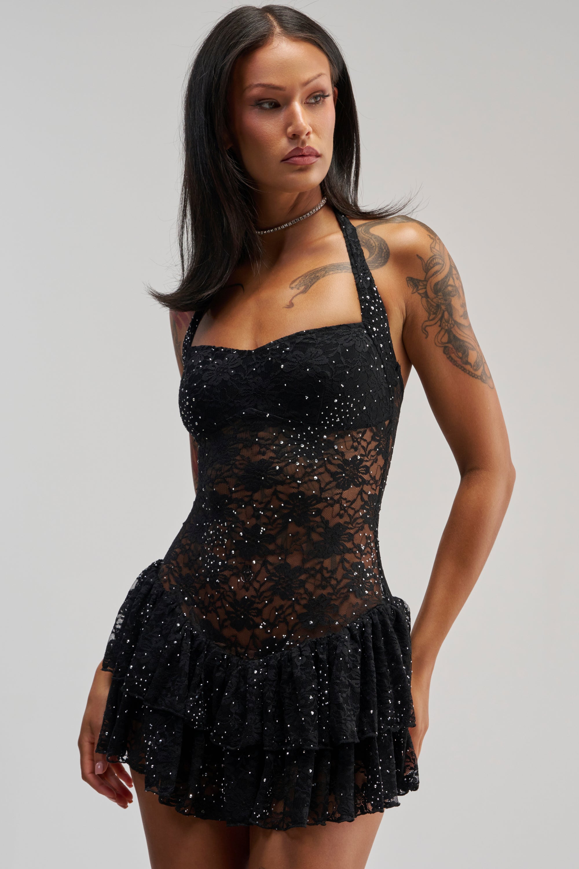 A woman with long dark hair wears the SEE RIGHT THROUGH ME LACE MINI DRESS IN BLACK, which features a sleeveless halter-neck design embellished with rhinestones and a ruffled skirt. Visible tattoos are on her arm as she gazes to the side against a plain background.