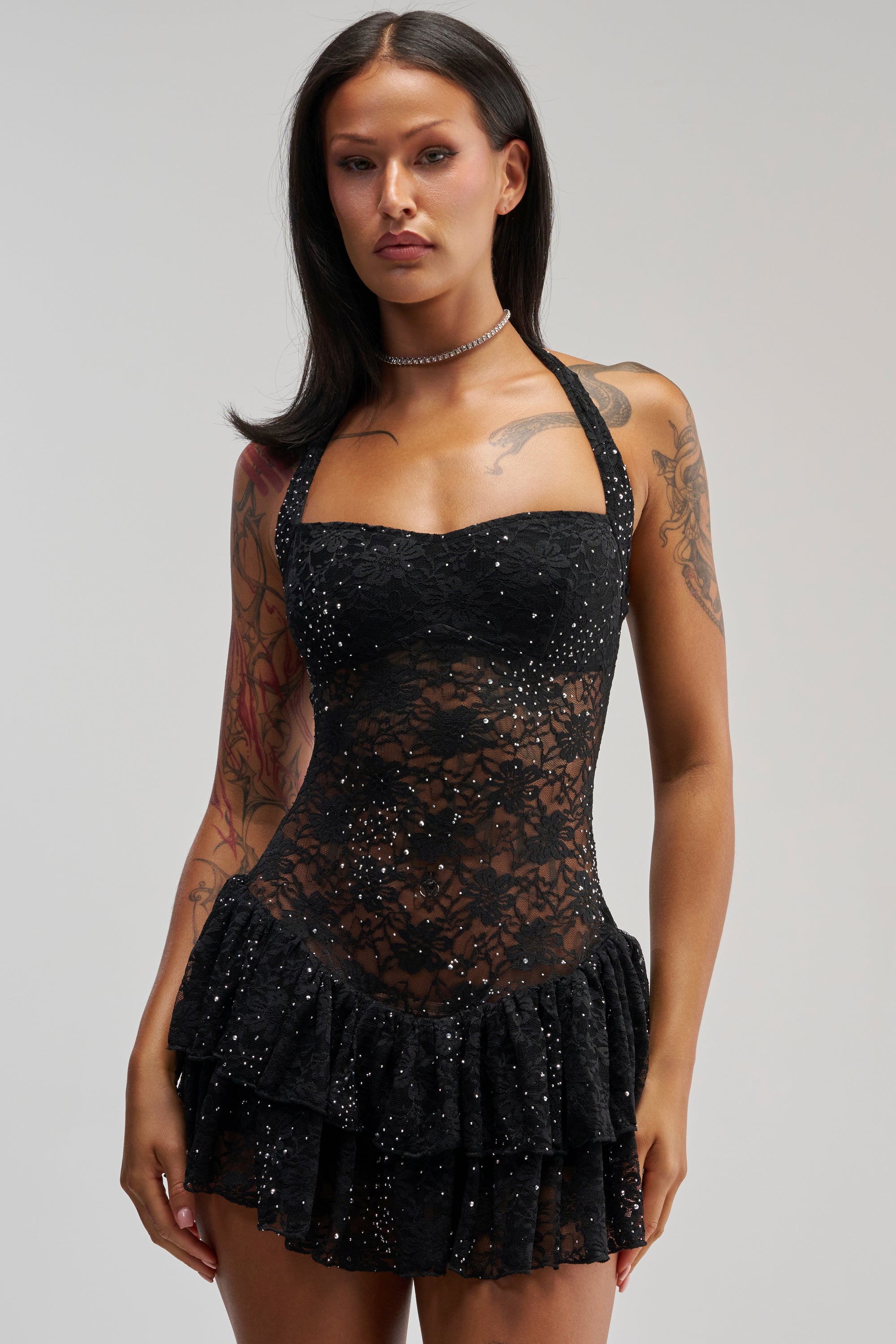 A woman with dark hair wears the SEE RIGHT THROUGH ME LACE MINI DRESS IN BLACK—a black lace mini dress with sheer, sparkly details and a ruffled skirt. She shows off her arm tattoos and accessorizes with a choker necklace while standing against a plain light background.