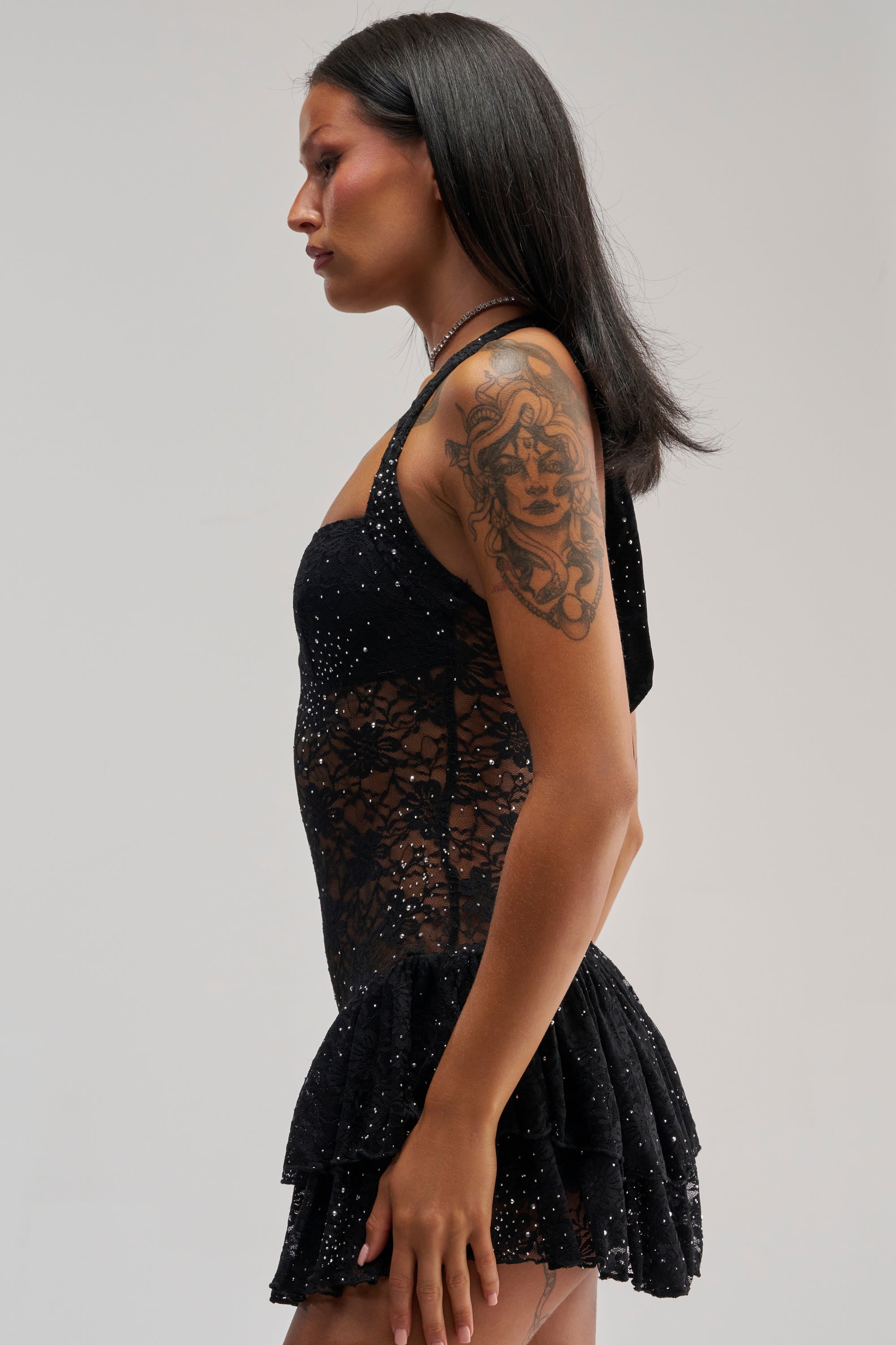 A person with long dark hair, wearing the SEE RIGHT THROUGH ME LACE MINI DRESS IN BLACK—adorned with sparkles—stands in profile. They have a visible tattoo on their upper arm and a neutral expression.