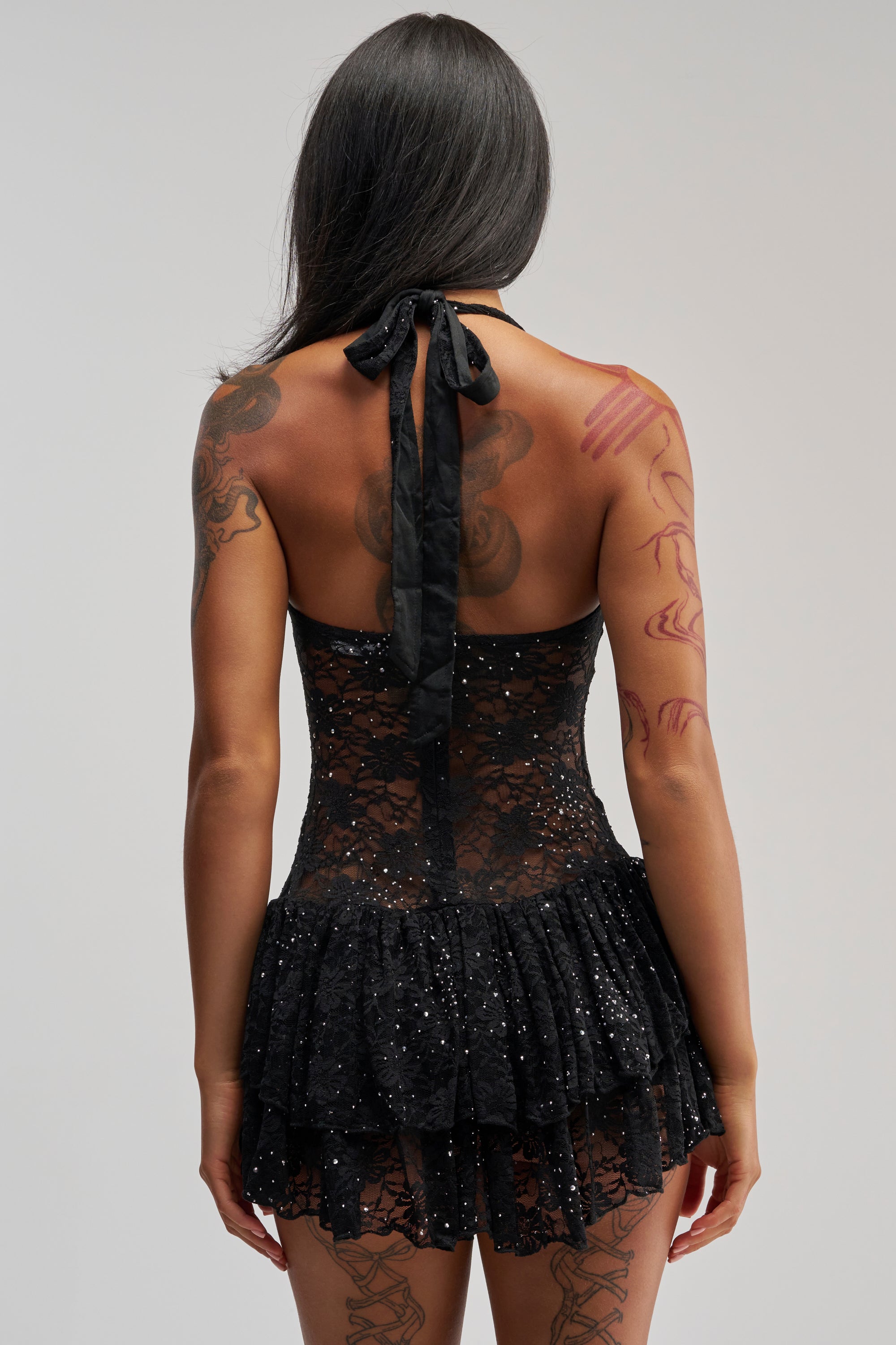 A person with long dark hair and visible tattoos is shown from the back, standing against a neutral background while wearing the SEE RIGHT THROUGH ME LACE MINI DRESS IN BLACK.