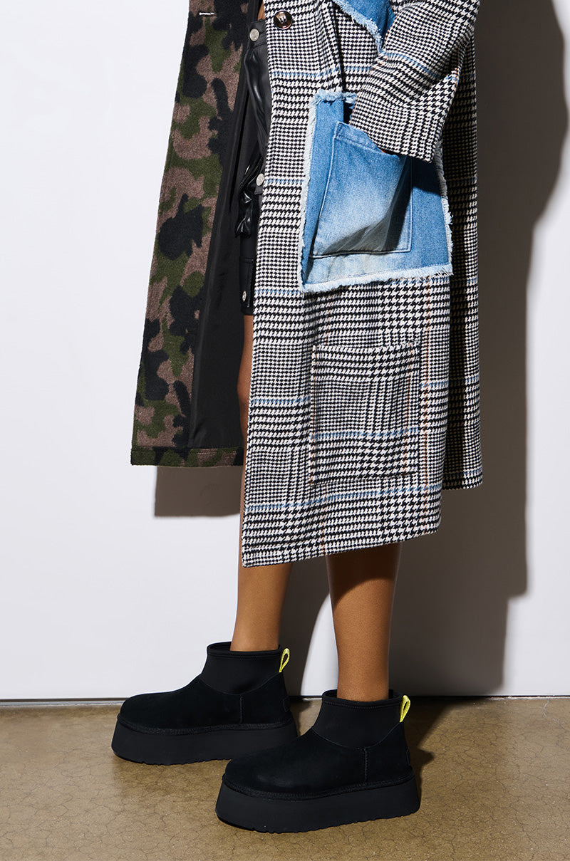 A person is showcasing a houndstooth and camouflage coat with a denim pocket detail, paired with the UGG CLASSIC MINI DIPPER IN BLACK boots. The emphasis is on their lower half against a white backdrop.