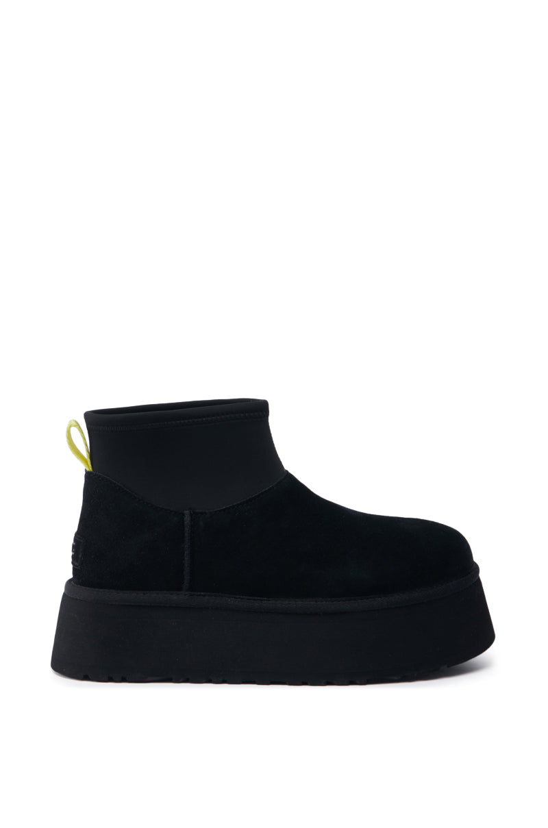 Introducing the UGG CLASSIC MINI DIPPER IN BLACK: This single black suede boot showcases a prominent thick platform sole. It features a neoprene-like upper collar complemented by a striking bright yellow pull loop at the back. Set against a white background, each distinctive design element is beautifully highlighted.