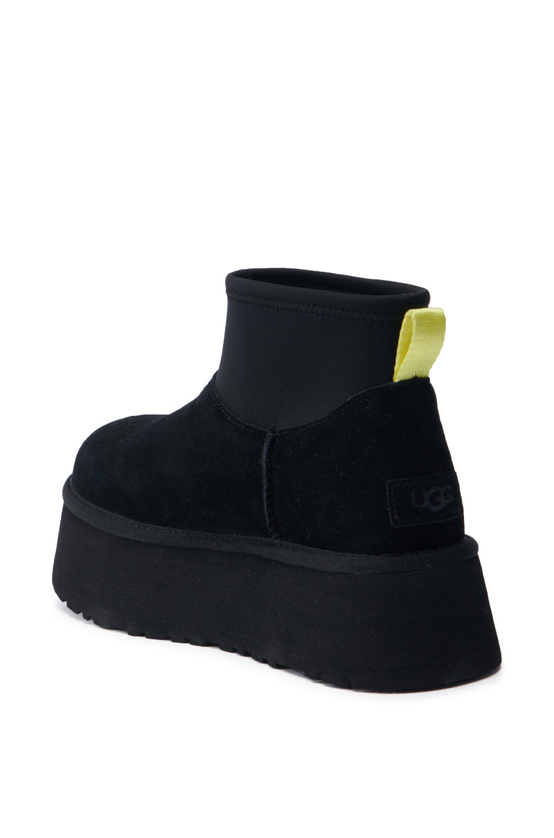 The UGG Classic Mini Dipper in Black showcases a chic suede upper as a black platform ankle boot, enhanced by a vibrant yellow pull tab at the back. The iconic UGG logo adorns the heel, and its chunky platform sole contributes to its modern appeal.