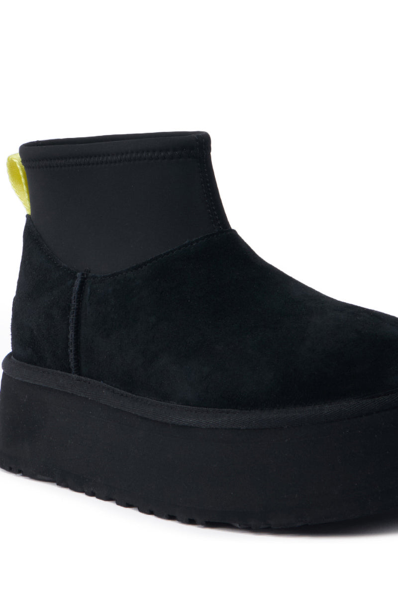 Introducing the UGG Classic Mini Dipper in Black, an ankle boot featuring black suede and a thick platform sole. This contemporary shoe is designed with a neoprene-like upper and an eye-catching yellow pull tab at the back, combining trendiness and style with every step.
