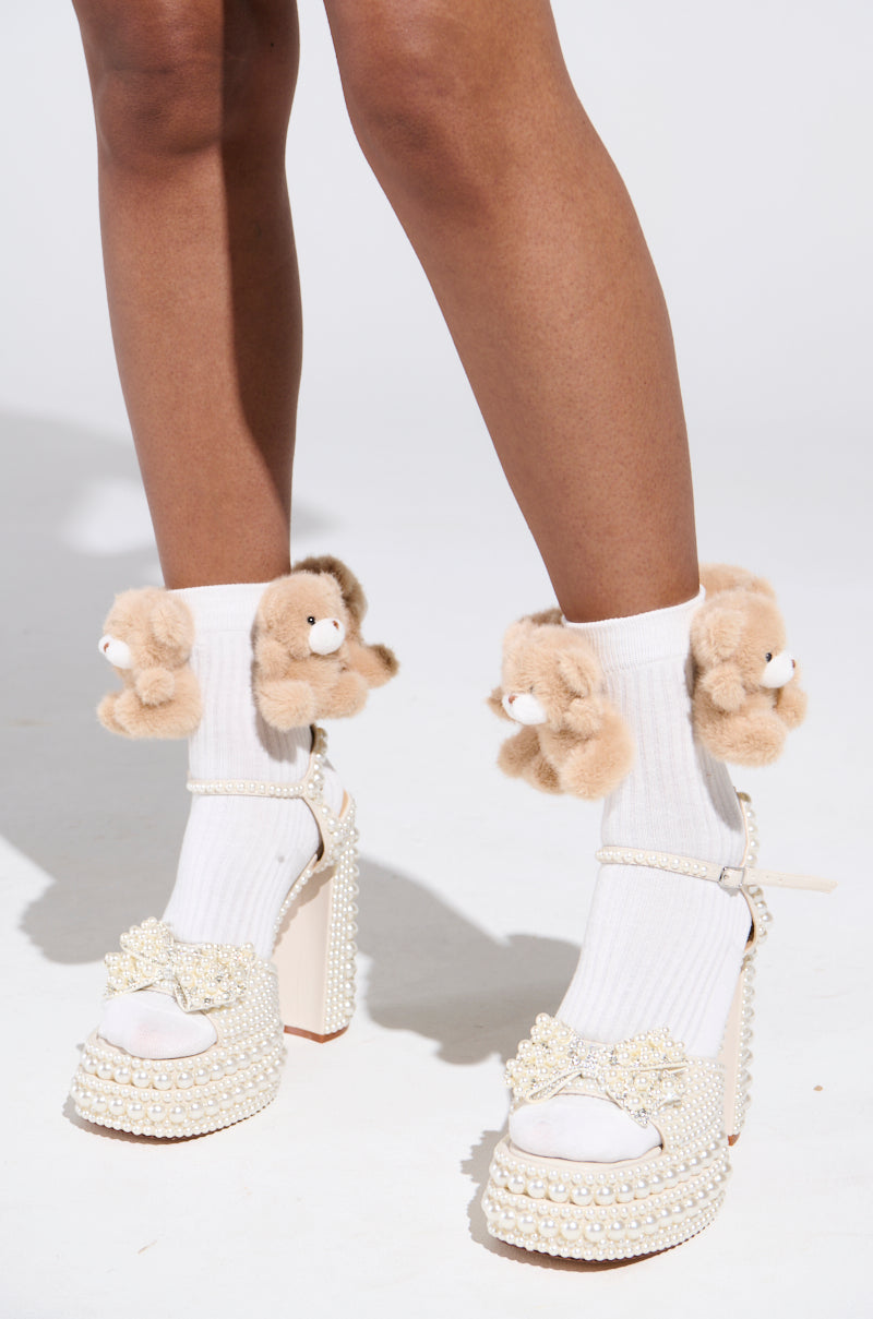 A close-up of the BEARY BABE SOCK displays white socks with plush tan bear appliques at the ankles, styled with pearl and bow embellished high heels. Made from stretchy material for comfort, it is showcased against a simple light gray background. 0