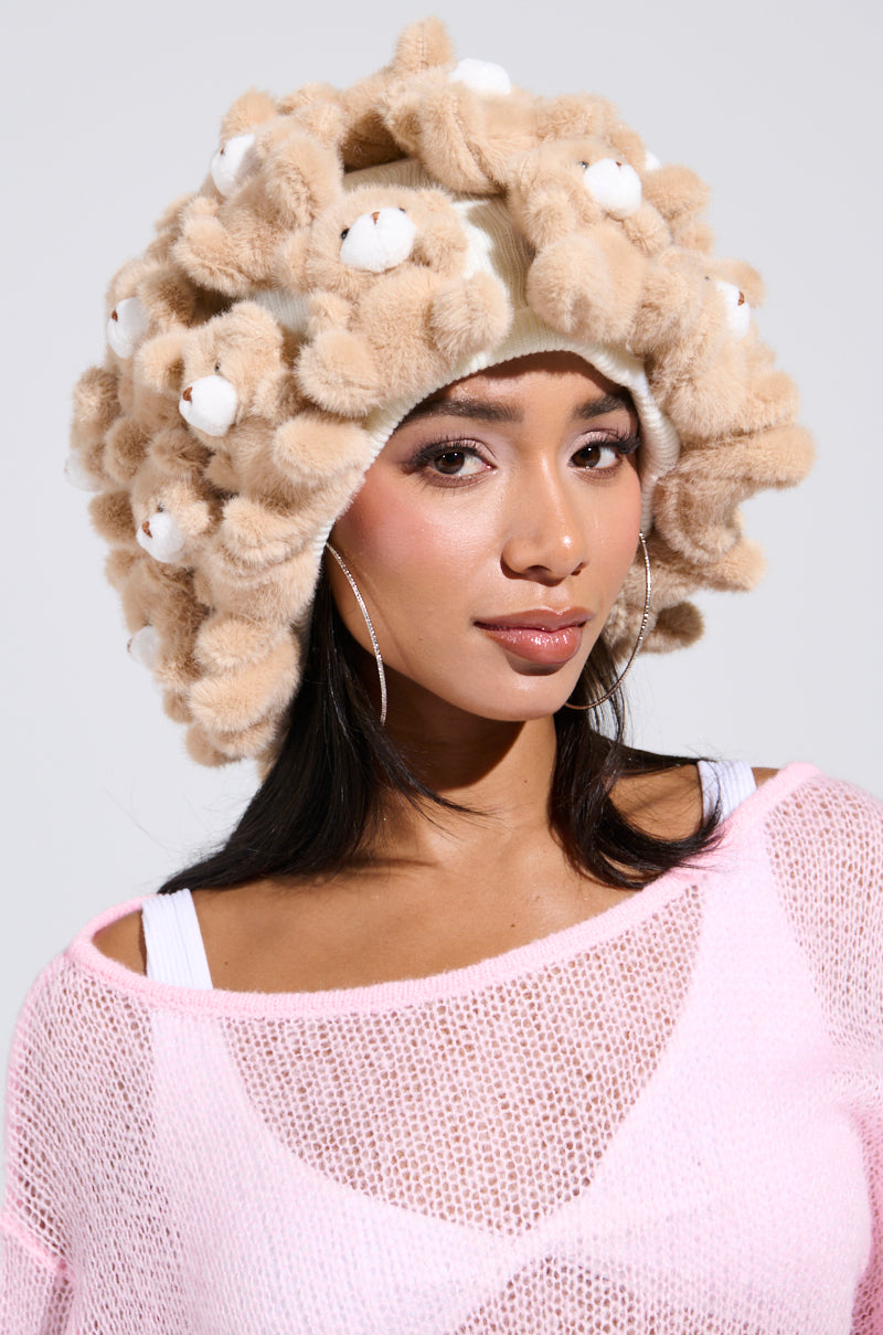 Wearing a BEARY BABE BEANIE adorned with fluffy bear appliques, a long-haired woman with hoop earrings smiles slightly against a plain background. Her ensemble's whimsical charm is enhanced by a pink mesh top layered over a white tank top. 0
