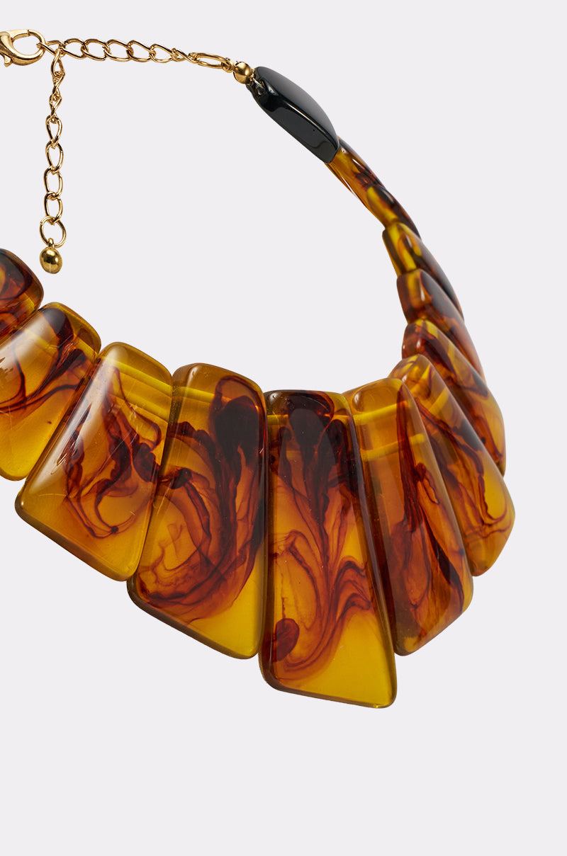A close-up of the JUST WANNA PARTY NECKLACE, a chunky resin statement piece with large, flat amber segments featuring dark swirls, a glossy finish, and an adjustable gold chain. 3