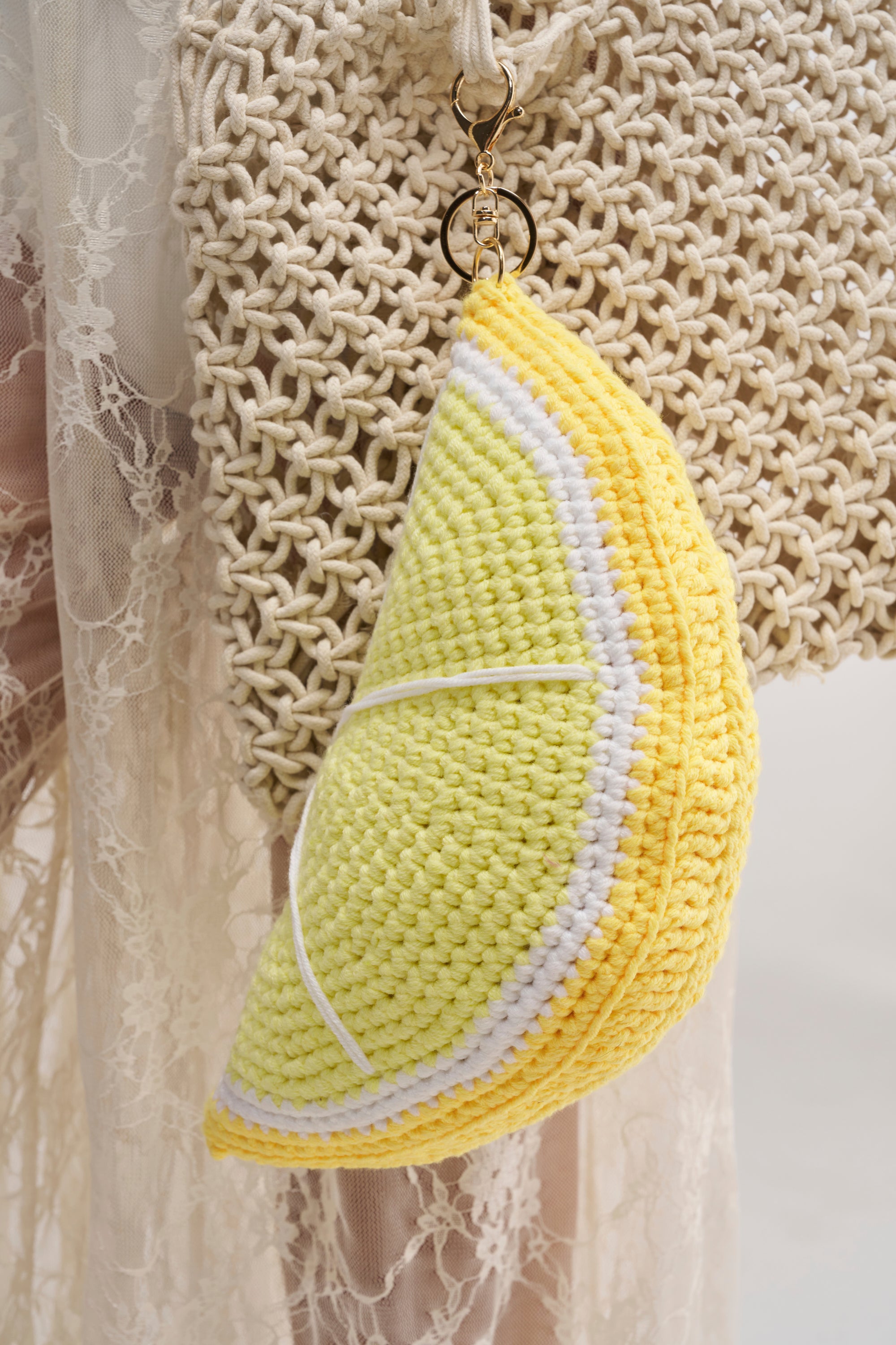 Close-up of the SO FRESH OVERSIZED CROCHET KEYCHAIN, a yellow and white lemon slice, hanging from a beige woven bag—a cute summer accessory or travel charm with lace fabric in the background.