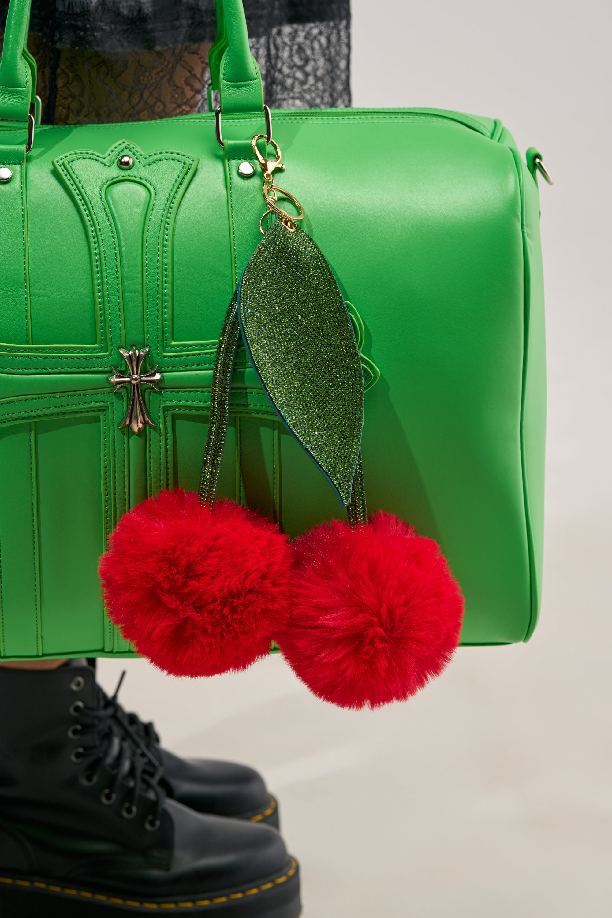 A bright green handbag with a metallic cross detail, accessorized with the POP IT OVERSIZED KEYCHAIN—featuring two large red faux fur pom-poms and a glittery green leaf charm—next to black lace-up boots.