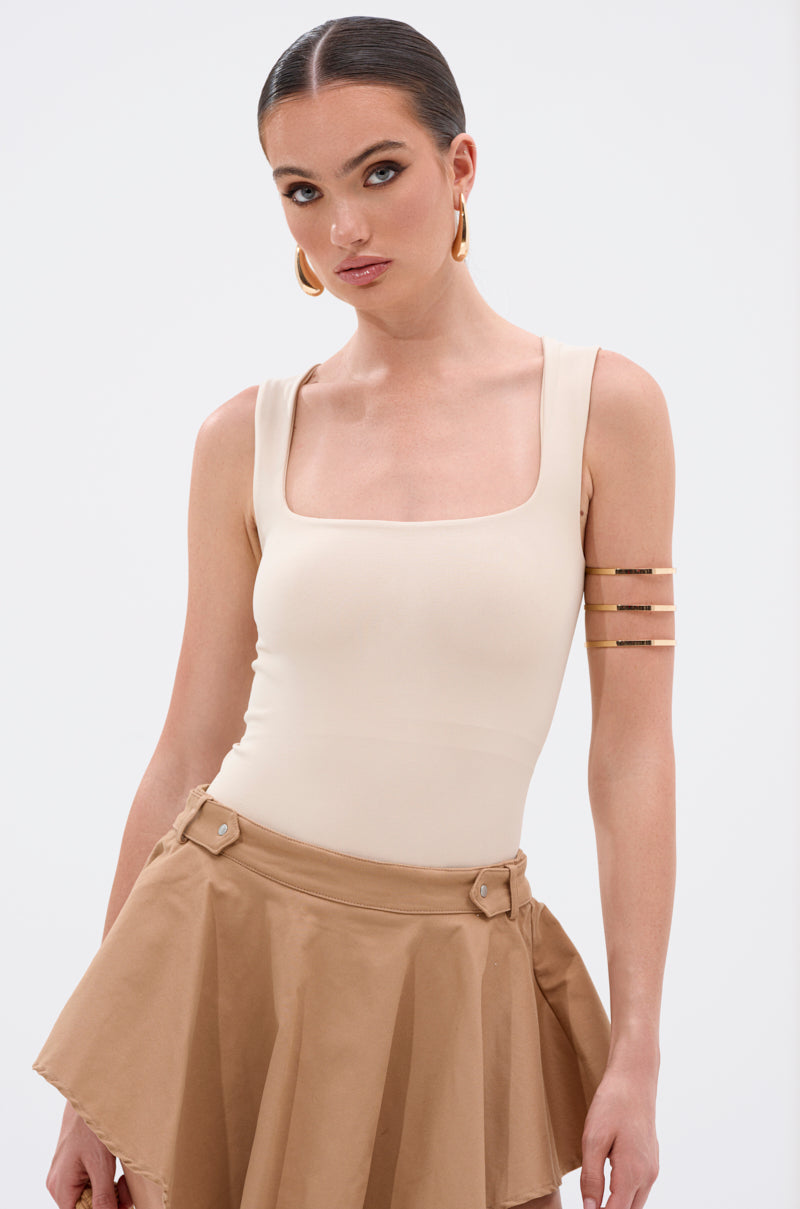 A woman with slicked-back hair models the NEW STANDARD SQUARE NECK SLEEVELESS BODYSUIT IN BEIGE, paired with a tan skirt and gold accessories. She stands against a plain white background, gazing slightly to the side.