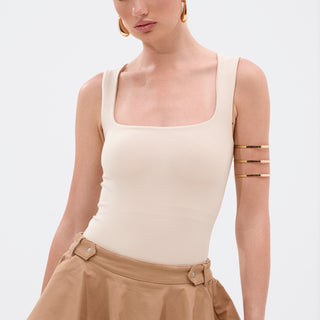 A woman with slicked-back hair models the NEW STANDARD SQUARE NECK SLEEVELESS BODYSUIT IN BEIGE, paired with a tan skirt and gold accessories. She stands against a plain white background, gazing slightly to the side.