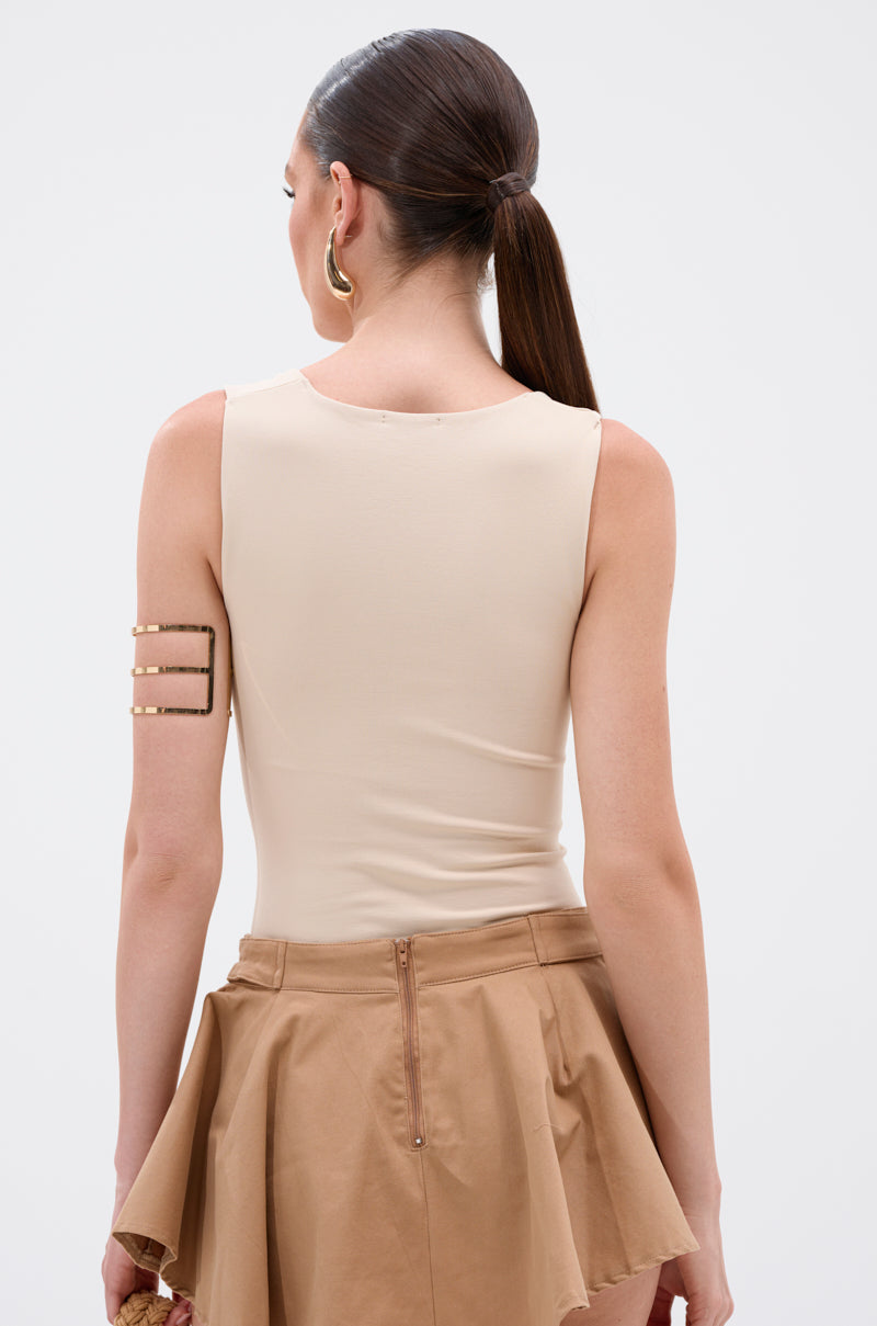 A woman with long brown hair in a low ponytail is seen from behind, wearing the NEW STANDARD SQUARE NECK SLEEVELESS BODYSUIT IN BEIGE, a tan zip-front skirt, hoop earrings, and a gold geometric arm cuff on her upper left arm. 3