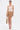 A woman poses against a white background in the NEW STANDARD SQUARE NECK SLEEVELESS BODYSUIT IN BEIGE, styled with a light brown mini skirt, strappy white heels, hoop earrings, slicked-back hair, and holding a woven handbag.