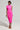 A person wearing the vibrant CAMARI Ruched Midi Dress in neon pink poses against a plain backdrop. They hold a small, sparkly clutch and wear brown strappy heels, with neatly styled hair adorned with earrings.