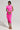 Against a plain backdrop, a woman showcases the CAMARI RUCHED MIDI DRESS, an off-the-shoulder evening attire in vibrant pink with elegant draping. She carries a chic clutch and dons strappy heels. Her hair is styled gracefully, complemented by her choice of earrings.