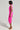 A person posed in profile against a simple backdrop, dressed in the vibrant CAMARI RUCHED MIDI DRESS paired with orange heeled sandals. They have short dark hair and are holding a small purse, emphasizing the chic style of this pink ruched piece.