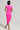 A person wearing the CAMARI RUCHED MIDI DRESS in a vibrant pink tone is positioned on a neutral background. The dress showcases gathered details and an asymmetrical hem. The individual has short, dark hair and is wearing strappy heels, posing from the back to highlight the chic design of the outfit.