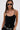 A woman with long brown hair wears narrow black sunglasses and the SEE THROUGH THE LINE CROCHET BODYSUIT, styled with high-waisted black shorts and a silver chain belt with decorative circular details, standing against a plain white background.