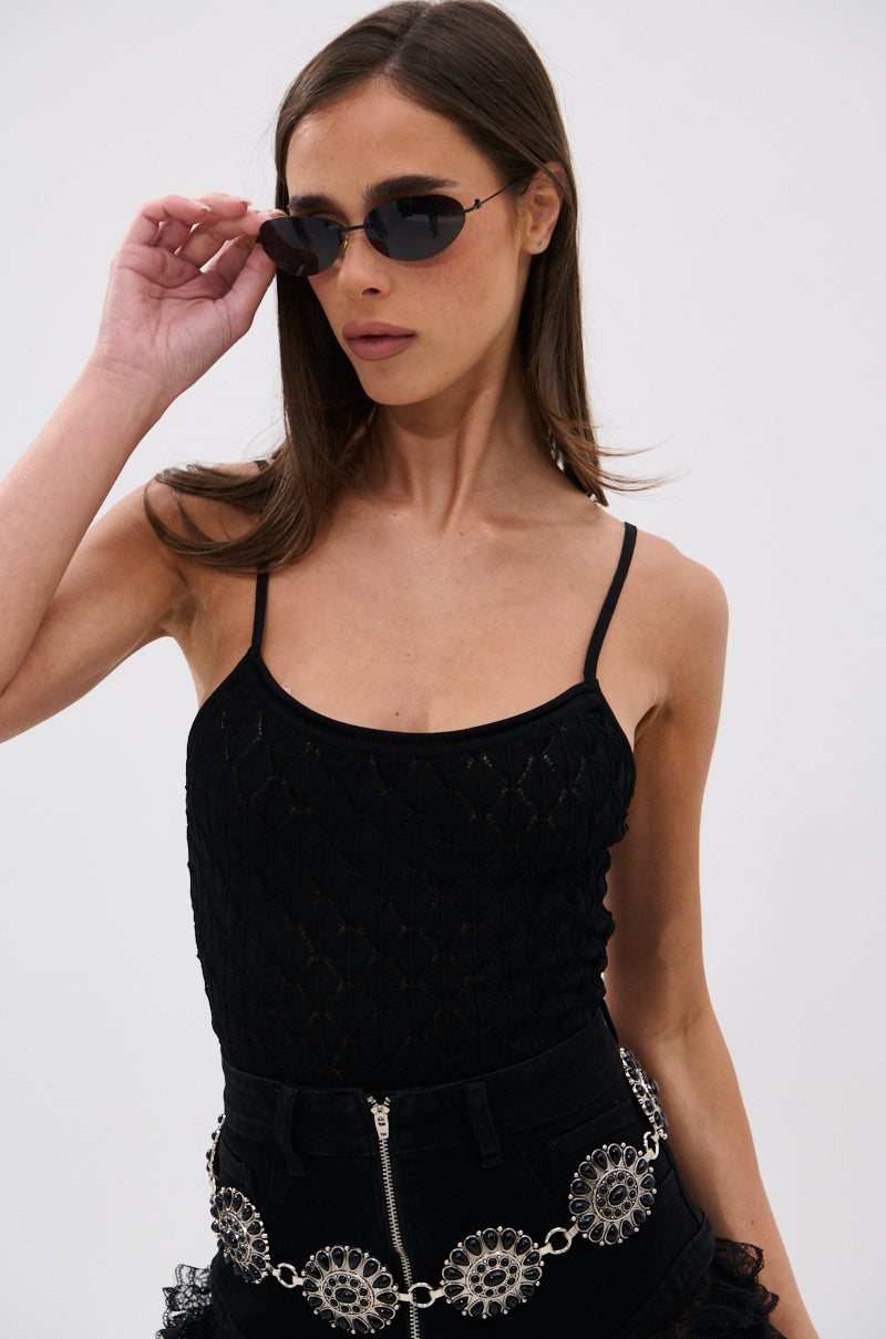 A woman poses against a white background wearing black sunglasses, black pants with a large silver belt, and the SEE THROUGH THE LINE CROCHET BODYSUIT featuring adjustable tank straps.