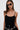 A woman with long brown hair poses against a white background, wearing black sunglasses, high-waisted black pants, a silver chain belt, and the SEE THROUGH THE LINE CROCHET BODYSUIT with adjustable tank straps.