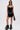 A woman wearing sunglasses, the SEE THROUGH THE LINE CROCHET BODYSUIT, high-waisted black fringe shorts, a silver chain belt, black fringe boots, and holding a black handbag stands against a plain white background.