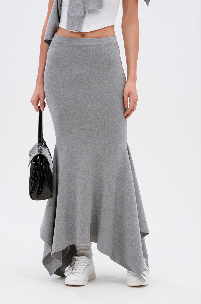 A woman wears the TAKE IT BACK KNIT MAXI SKIRT in gray with a white top, gray sweatshirt tied around her shoulders, white sneakers, and holds a black handbag. The background is plain white.