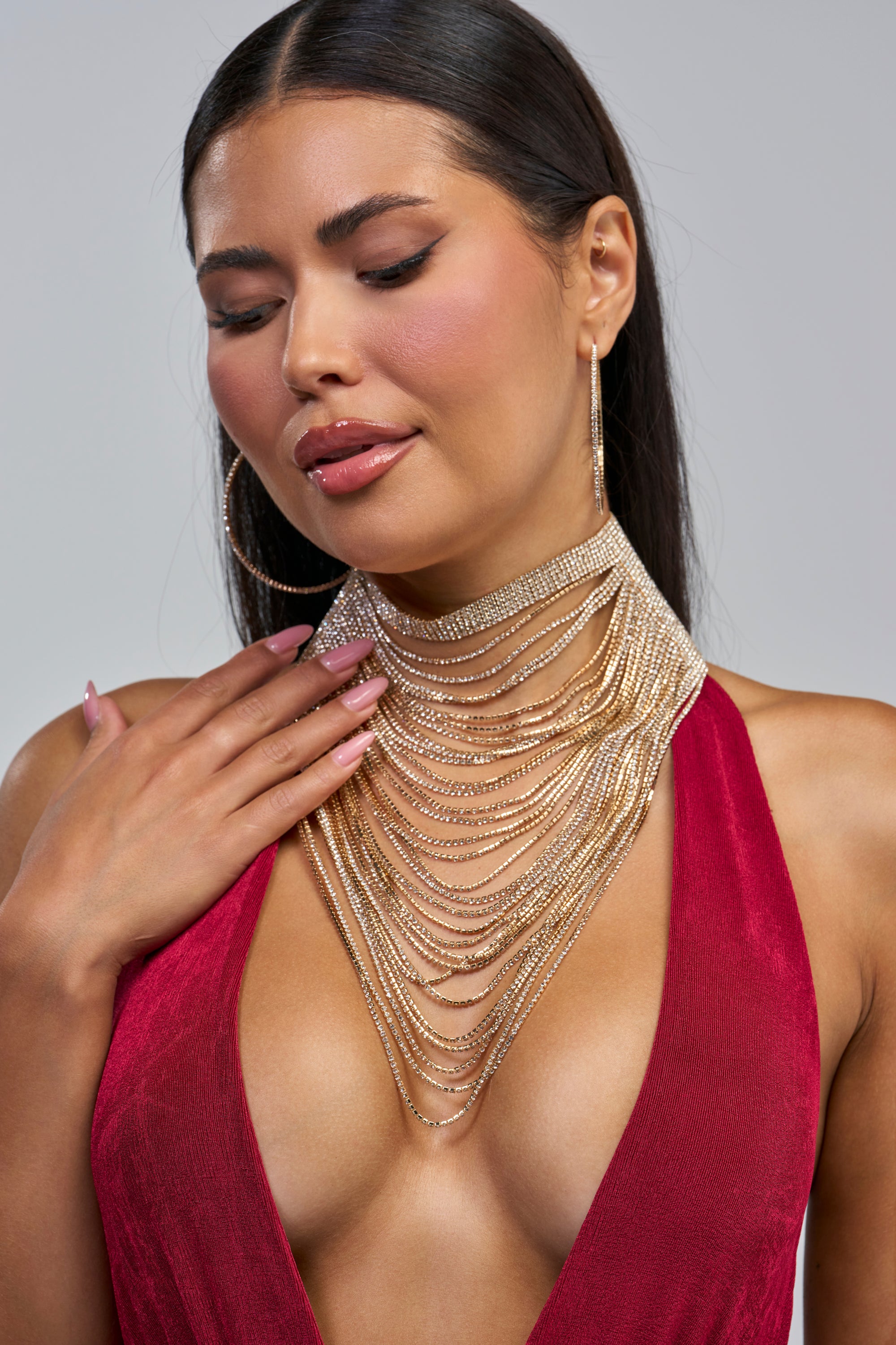 Woman with long dark hair wearing a deep red dress, large hoop earrings, and the SHES A FOX NECKLACE—a layered gold chain statement piece paired with a delicate gold choker—looking down with one hand touching her chest.