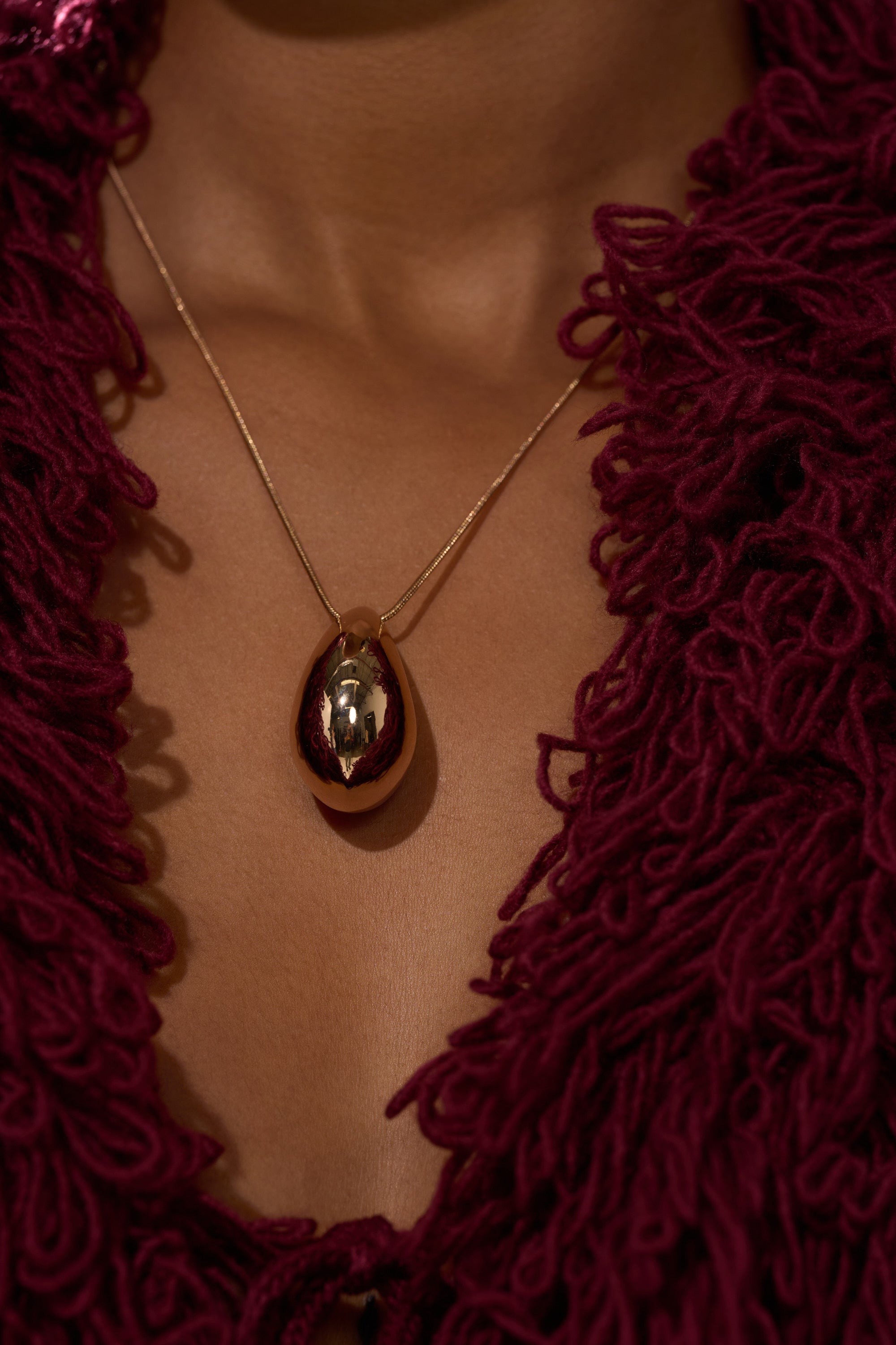 A close-up of a person wearing the DESIGNER DREAMING NECKLACE—a gold teardrop pendant on a rope chain—paired with a deep burgundy textured garment with thick, looped yarn details. The necklace and rich fabric texture are in focus.