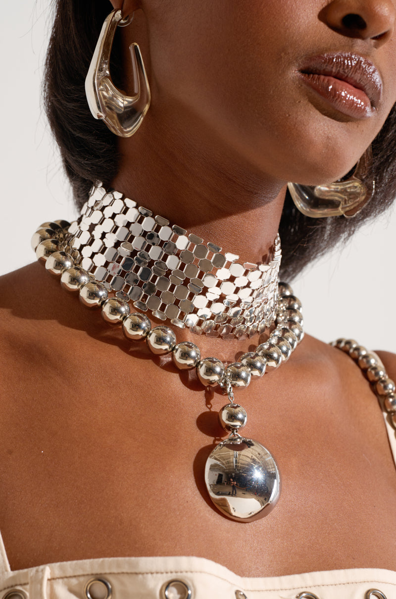 The image displays a person wearing the POP BUBBLY NECKLACE SET, including large silver statement earrings and a layered silver choker necklace with metallic mesh and beaded layers. The reflective pendant complements the medium-dark skin and cream-colored top beautifully.