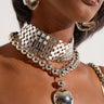 Close-up of a person wearing a shiny, metallic jewelry set. The set includes large hoop earrings, a thick choker with a honeycomb pattern, and the POP BUBBLY NECKLACE featuring circular bead detailing and a reflective pendant. The person's skin is medium-brown, and they are wearing a beige top.