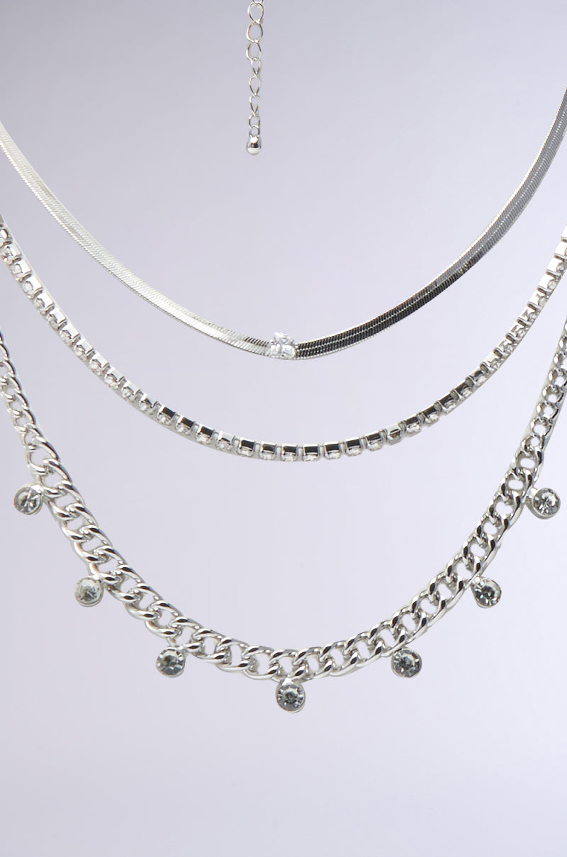 The LAYERED UP SILVER NECKLACE features three silver chains—a flat herringbone, a box chain, and a chunky chain with round charms—layered together against a light gray background for a stylish look. 1