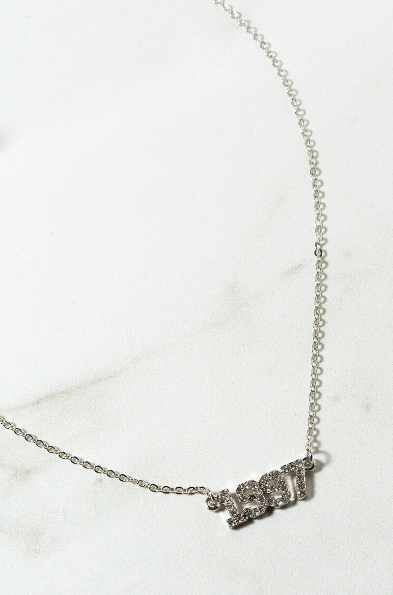 A close-up image of the delicate 2000 Rhinestone Year Necklace. The necklace features a pendant that spells out "2000," with each digit adorned with small, sparkling crystals. The fine chain sits on a light, neutral background.