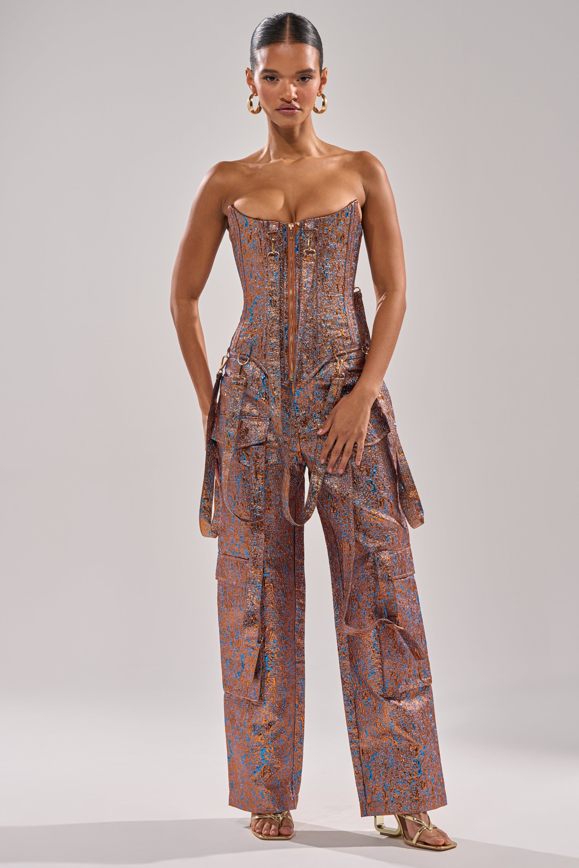 A woman faces forward in the CHECK IT OUT BROCADE BRONZE TROUSER, styled with a fitted strapless metallic top, wide-leg cargo details, slicked-back hair, gold hoop earrings, and strappy gold heels. 0