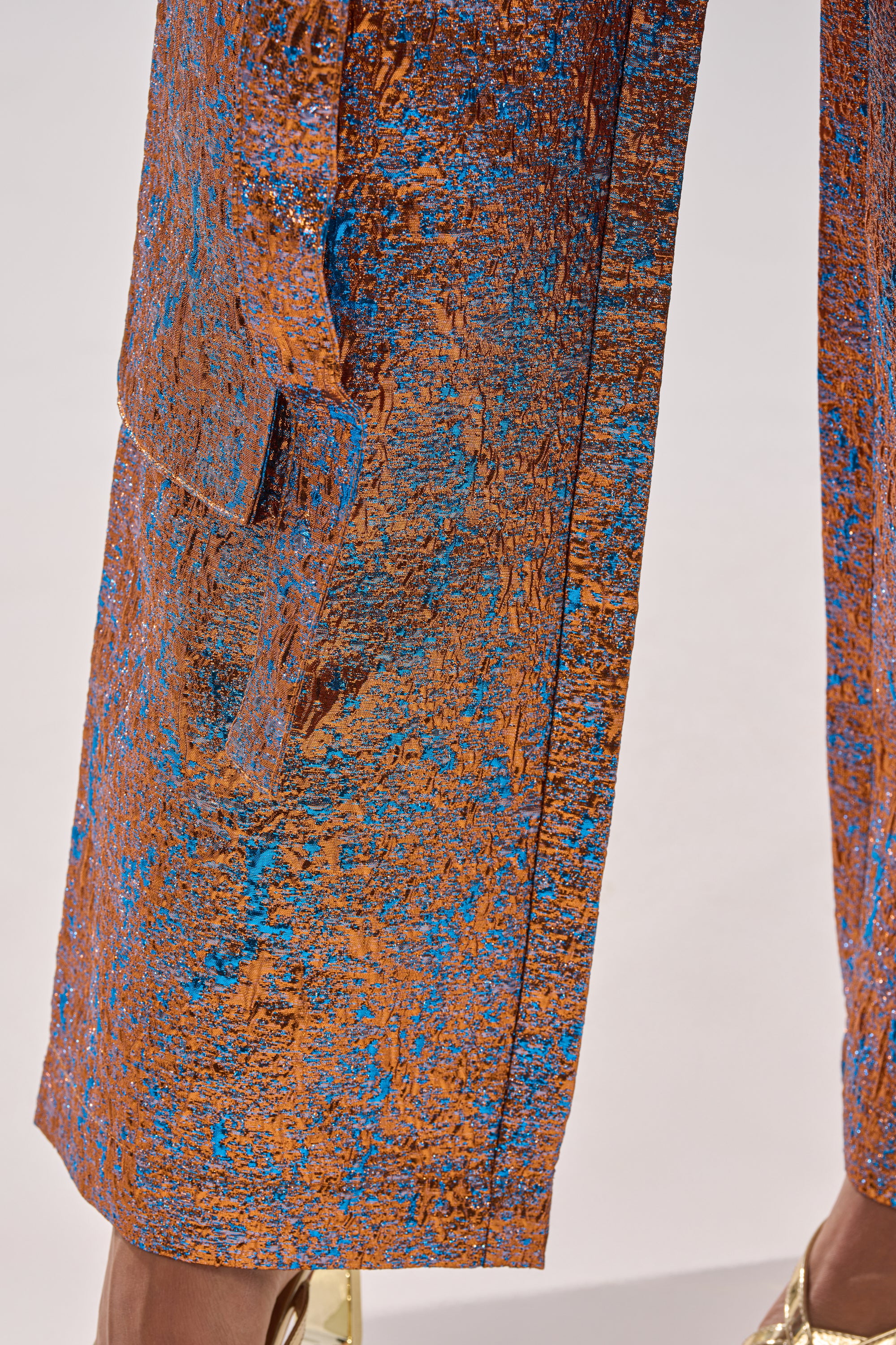 Close-up of CHECK IT OUT BROCADE BRONZE TROUSER pant legs in coppery textured metallic fabric with blue accents and flap pocket detail, paired with shiny gold shoes, set against a light background. 10
