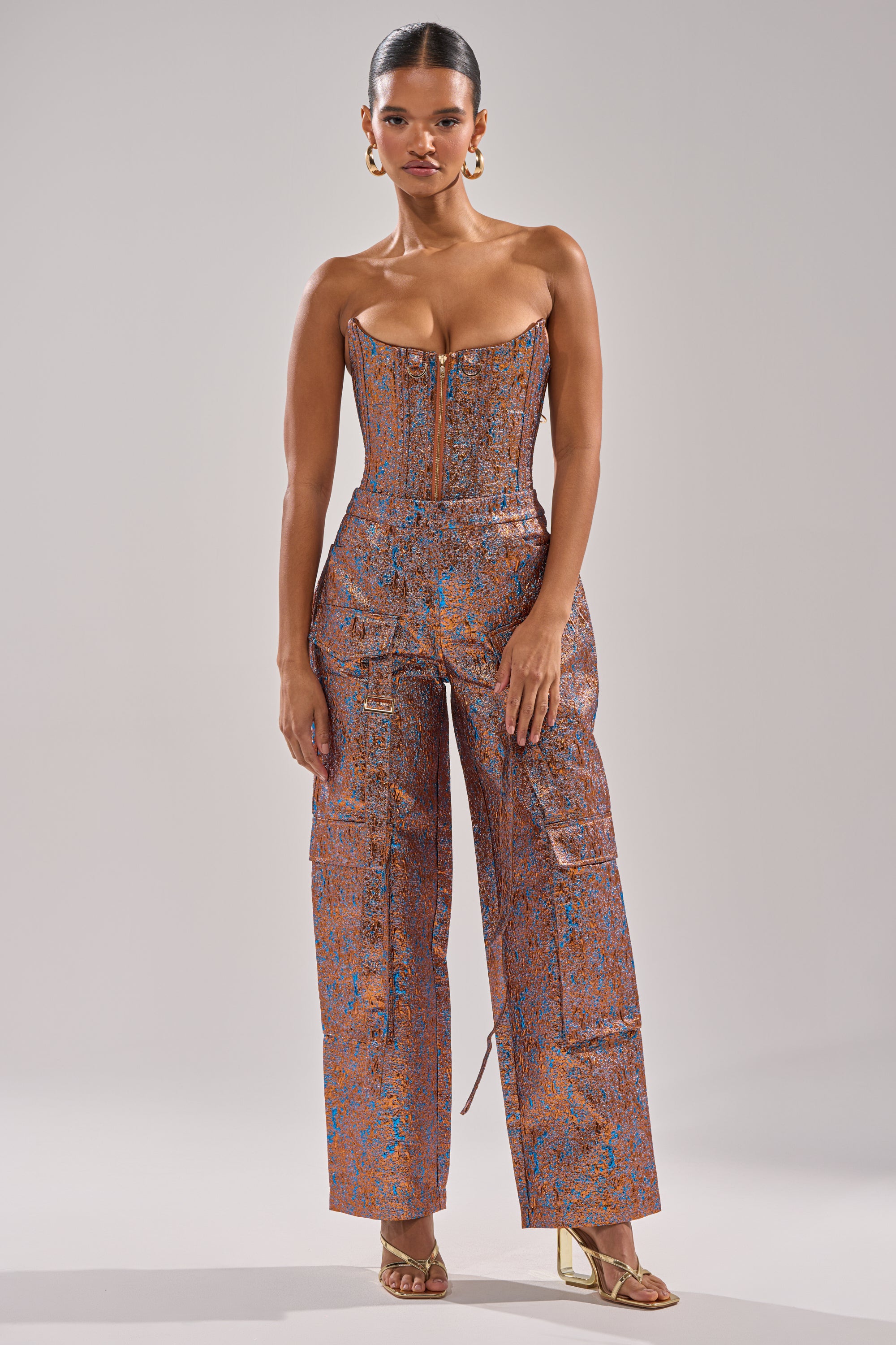 A woman stands facing forward in the CHECK IT OUT BROCADE BRONZE TROUSER with a fitted bodice and wide-leg metallic pants. She has slicked-back hair, gold hoop earrings, and gold heeled sandals against a plain light gray background. 1