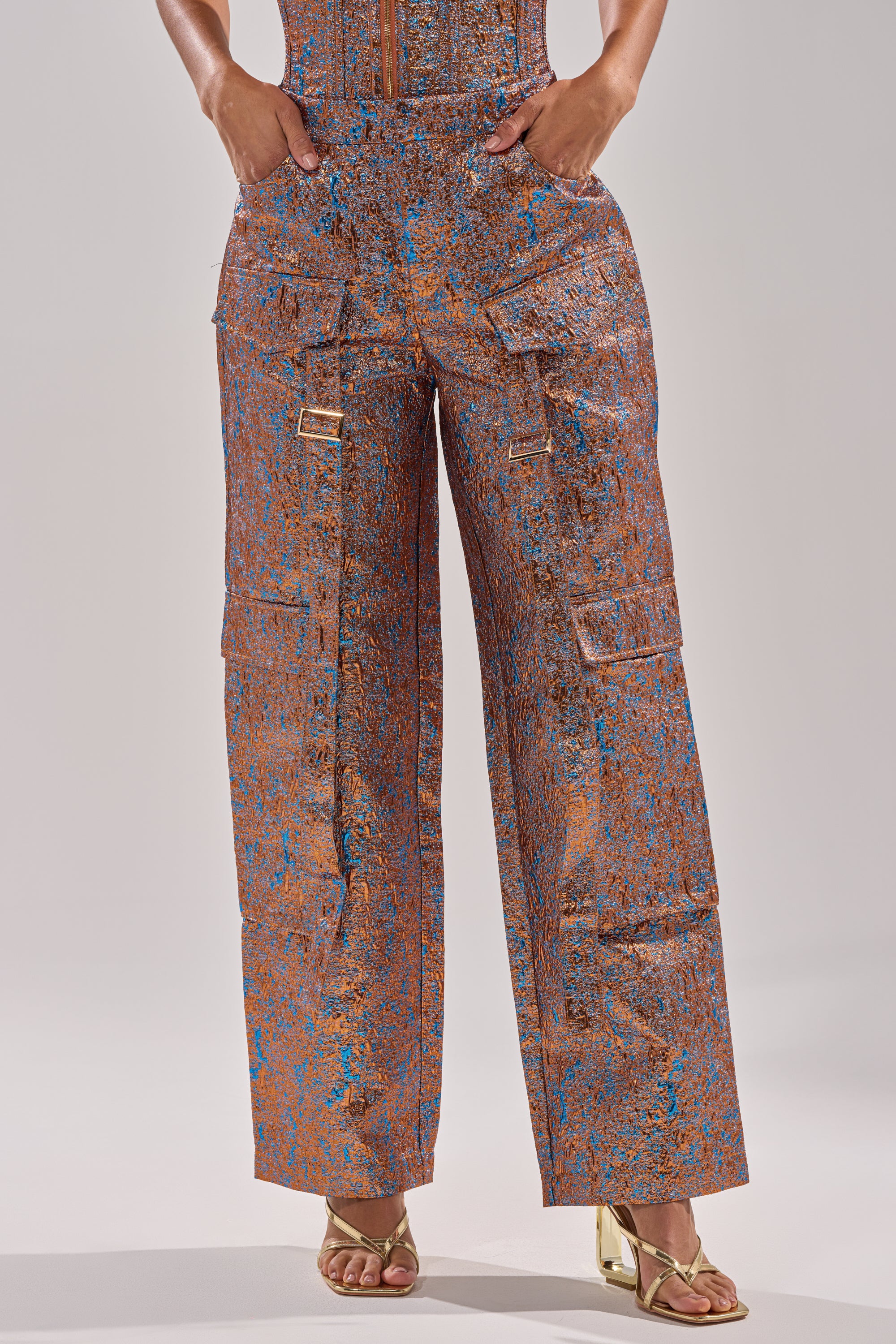 Wearing the CHECK IT OUT BROCADE BRONZE TROUSER featuring metallic blue and copper patterns, a person stands in gold open-toe heels. Only their lower torso and legs appear against a plain backdrop. 2