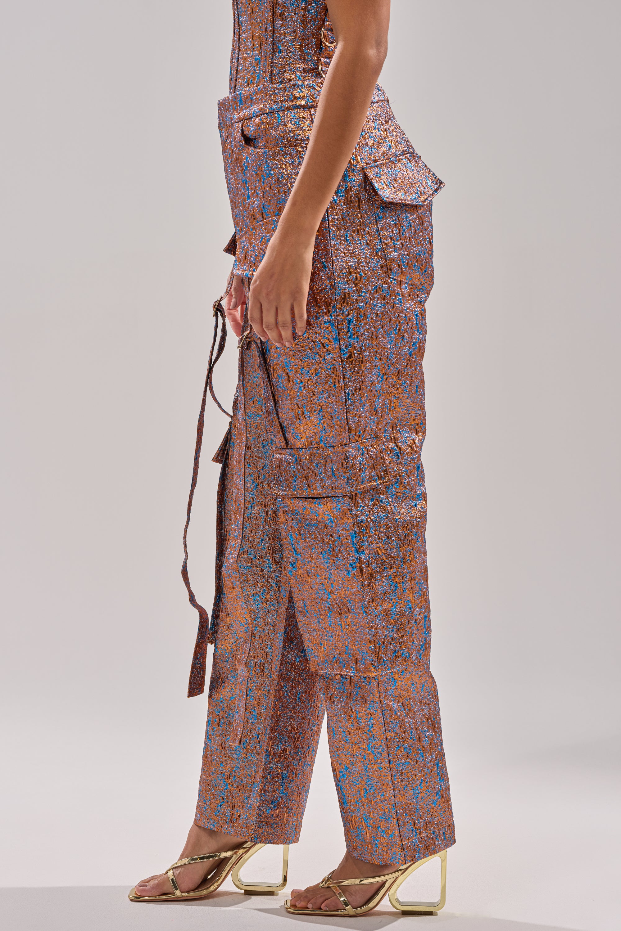 A person in profile wears the CHECK IT OUT BROCADE BRONZE TROUSER—shimmering metallic orange-blue wide-leg pants with side pockets and a belted waist—paired with gold heeled sandals, set against a plain light background. 3