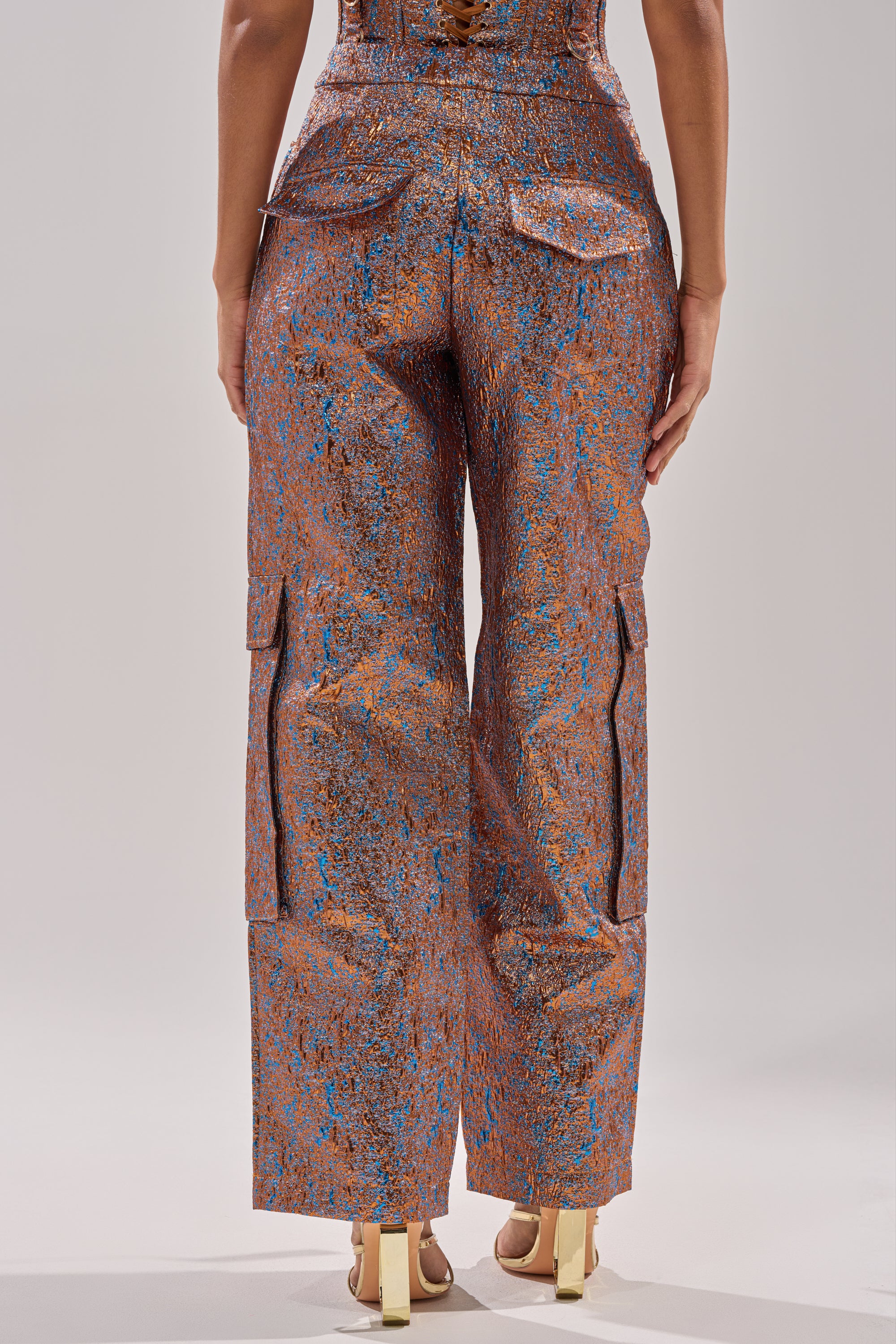 A person stands facing away, wearing the CHECK IT OUT BROCADE BRONZE TROUSER—high-rise, straight-leg metallic pants with blue accents and large back pockets—styled with gold heels against a plain, light backdrop. 4