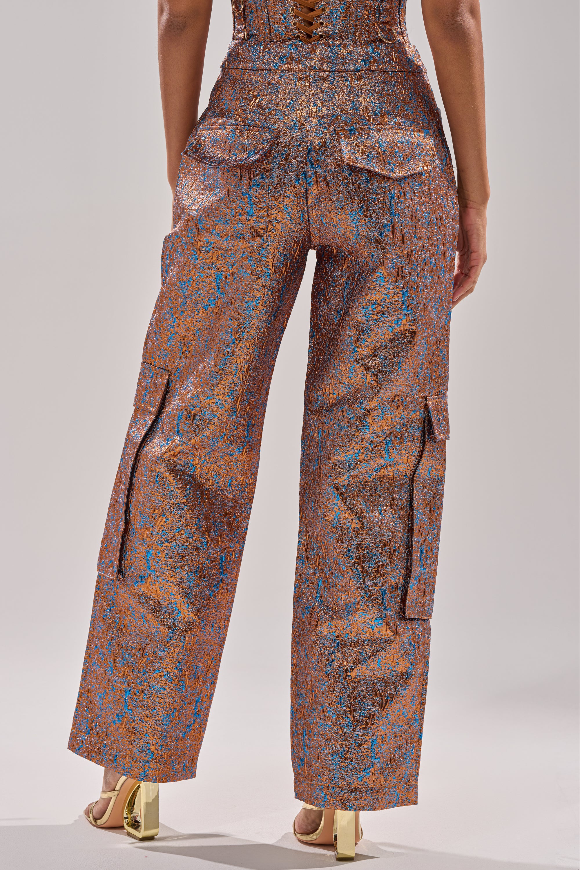 A person stands facing away, wearing the CHECK IT OUT BROCADE BRONZE TROUSER with a copper and blue textured pattern, high-rise straight-leg fit, back flap pockets, side cargo pockets, and gold high-heeled sandals. 5