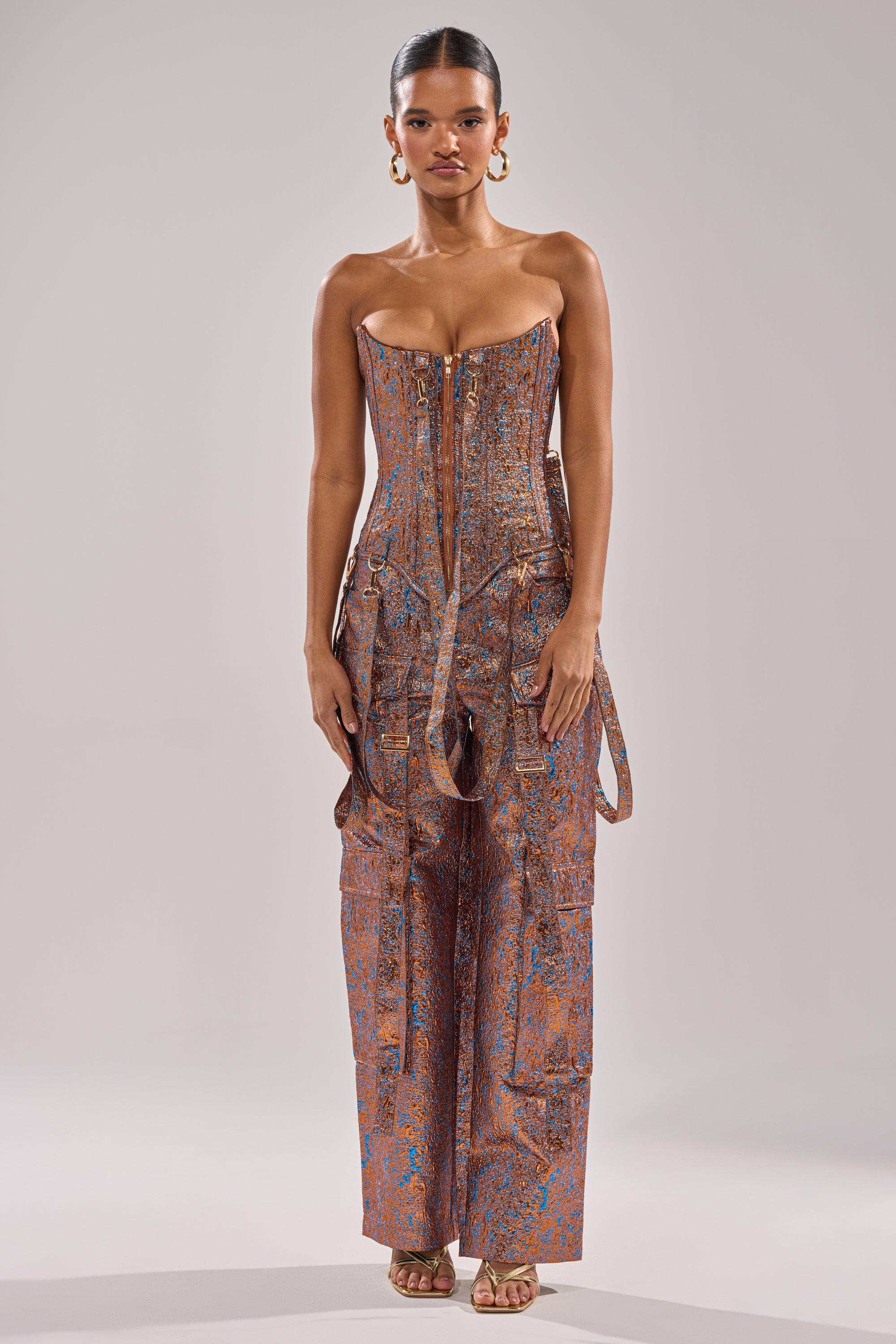 A woman faces forward in the CHECK IT OUT BROCADE BRONZE TROUSER, a strapless fitted jumpsuit with wide metallic pants, bronze brocade effect, hanging straps, gold earrings, and high-heeled sandals against a light gray background. 7