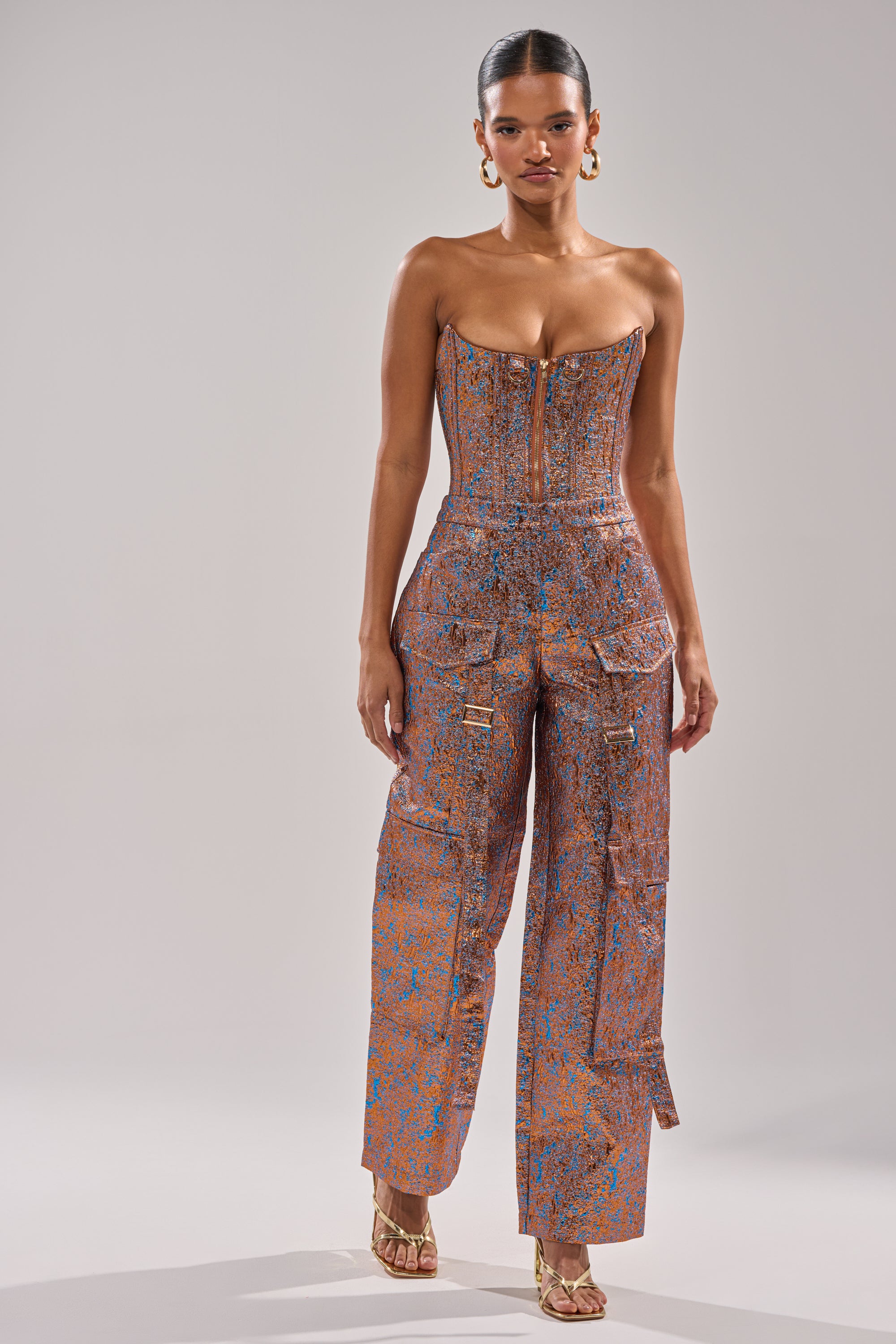 A woman poses against a neutral background wearing the CHECK IT OUT BROCADE BRONZE TROUSER by AKIRA Label, styled as a strapless fitted jumpsuit with wide legs and large pockets. She accessorizes with hoop earrings and gold heels, hair pulled back. 8