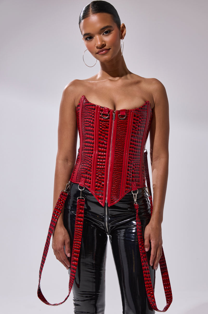A woman models the OH YEAH CORSET, a strapless red piece with buckle details, paired with black patent leather pants and hoop earrings. Her sleek hair is pulled back as she stands against a plain light background.