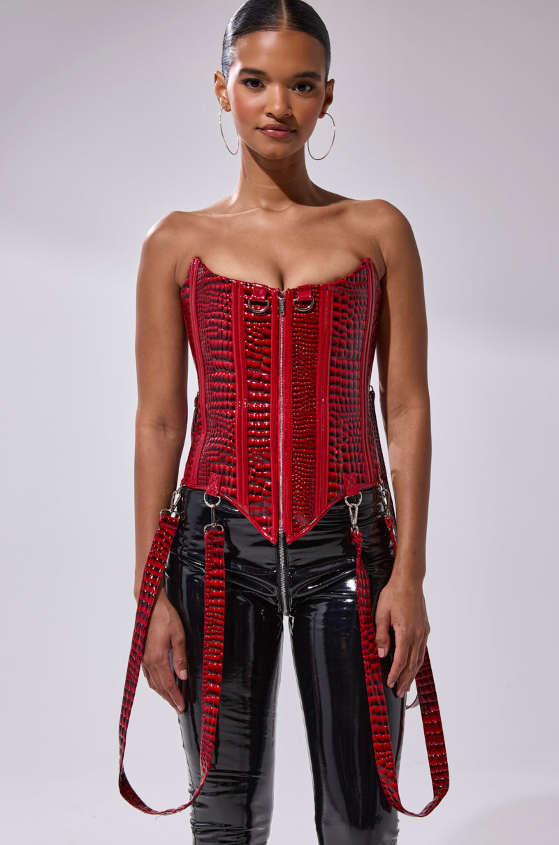 A woman faces forward in the OH YEAH CORSET, featuring red faux crocodile leather, zipper details, silver hardware, and detachable garter straps. She pairs it with shiny black pants, hoop earrings, and slicked-back hair.