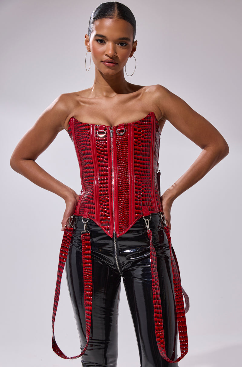 A woman stands confidently against a plain background, hands on hips, wearing the OH YEAH CORSET in shiny red faux crocodile leather with zipper details, paired with black patent leather pants.