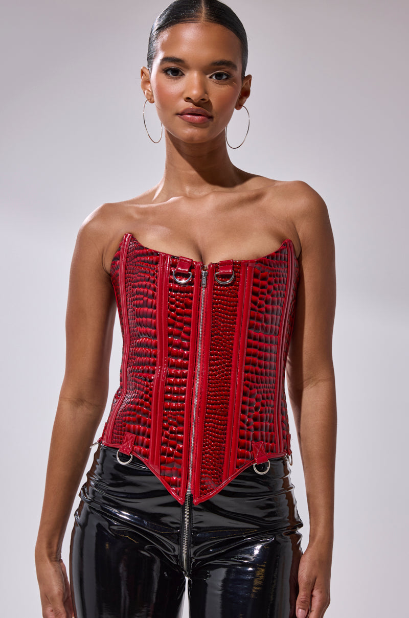 A woman wears the OH YEAH CORSET in shiny red faux crocodile leather with a front zipper, metallic details, and detachable garter straps, paired with glossy black pants against a neutral background.