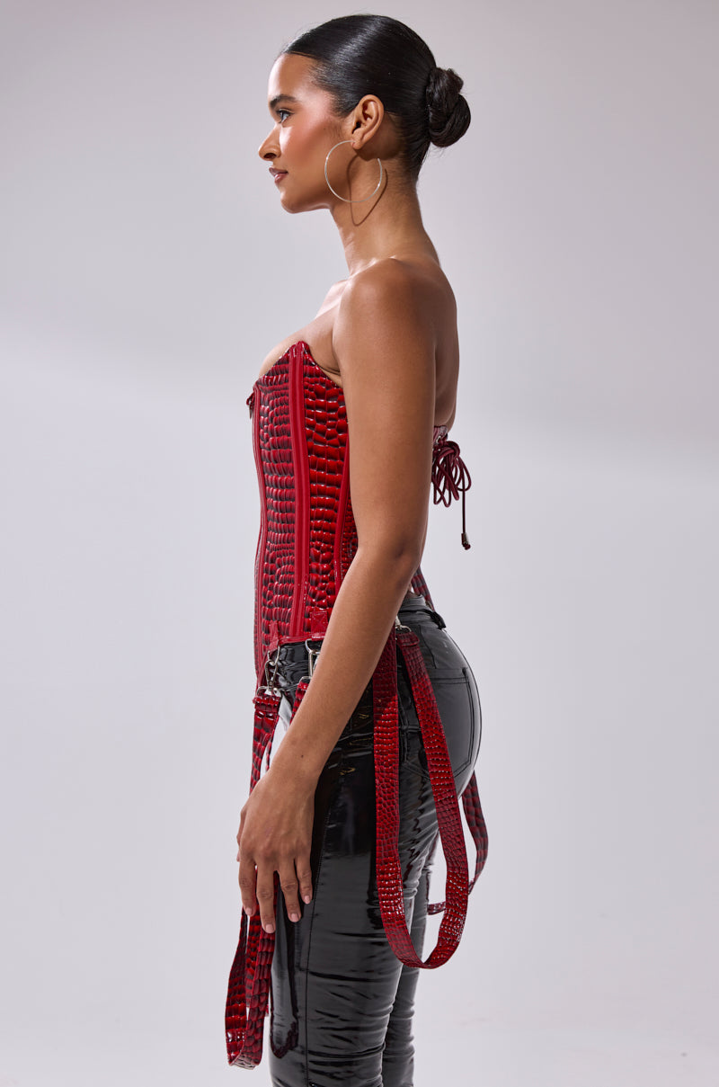 A woman stands in profile against a plain background, wearing the OH YEAH CORSET—a red strapless piece with an adjustable lace-up back—paired with shiny black high-waisted pants. Her hair is in a neat bun and she wears large hoop earrings.