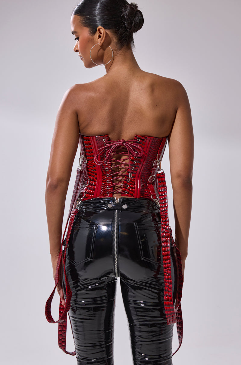 A woman faces away, wearing shiny black vinyl pants and the OH YEAH CORSET in red faux crocodile leather with detachable garter straps. Her hair is in a sleek bun, and she sports large hoop earrings against a plain light background.