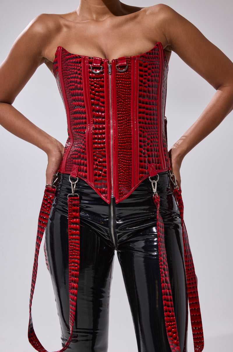 A person wearing the OH YEAH CORSET in red faux crocodile leather with metal hardware and black shiny vinyl pants stands with hands on hips against a plain background.
