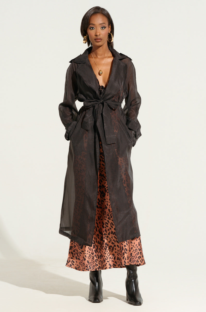 A woman stands confidently, showcasing an autumnal layering look with the CHIFFON CHATEAU LONG TRENCH IN BLACK tied at the waist over a leopard print dress. She accessorizes with gold earrings, a necklace, and black ankle boots. With a calm expression and hands in her coat pockets, she exudes a femme fatale finish against a light background.